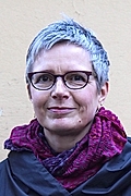 A woman with short gray hair and glasses, wearing a dark coat and a vibrant purple scarf, smiling against a plain background.