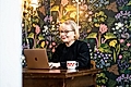 A woman in glasses works on a laptop at a wooden table, with a floral wallpaper background and a coffee mug.