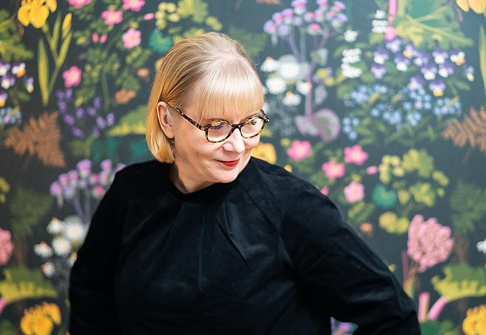A woman with short blonde hair and glasses poses against a colorful floral backdrop, wearing a black top.