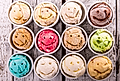 Twelve ice cream scoops in cups, each with a smiling face design, arranged on a rustic wooden surface.