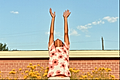 A child stands in a field of yellow flowers, raising arms towards a blue sky with wispy clouds.
