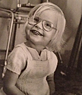 Black and white photo of a smiling young child with short blonde hair and large glasses, sitting indoors.
