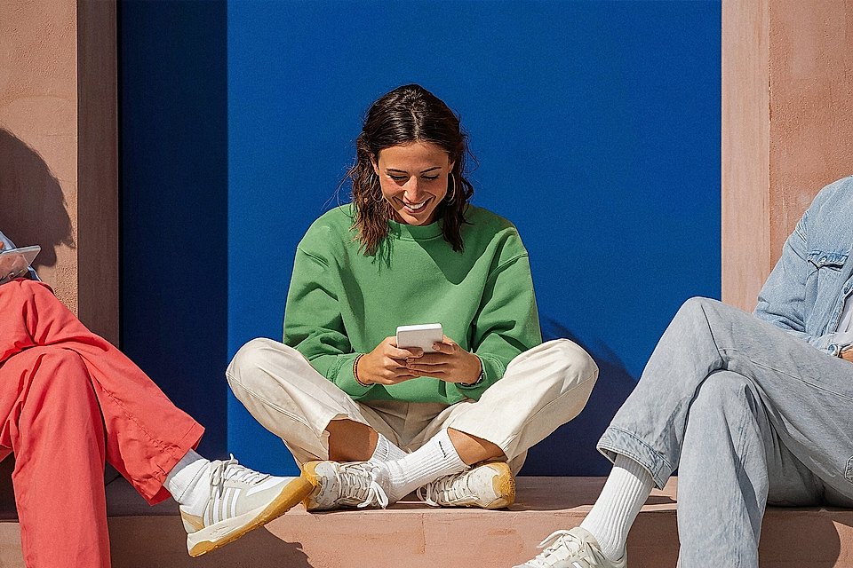 Three casually dressed young adults sit outdoors on steps, one smiling while using a smartphone.