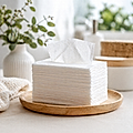 A stack of white tissue paper on a wooden plate, with a decorative vase and soft cloth in the background.