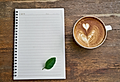 A blank notepad, a green leaf, and a latte with heart-shaped foam on a wooden surface.