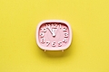 A pink square alarm clock showing the time 10:10 on a bright yellow background.