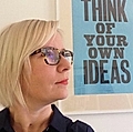 A woman with short blonde hair and glasses poses beside a framed sign that reads 'THINK OF YOUR OWN IDEAS'.