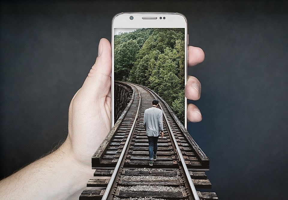 A hand holds a smartphone displaying a person walking on railway tracks in a forested landscape.