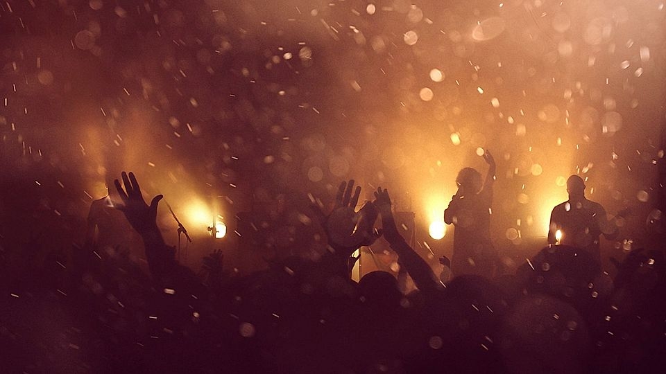 Silhouetted concert crowd with raised hands under warm lighting, creating a lively atmosphere.