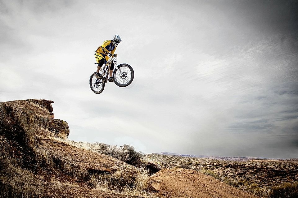 A mountain biker in yellow jumps off a rocky cliff, performing a stunt in a scenic outdoor landscape.