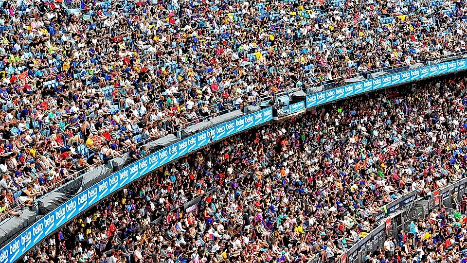 A large crowd fills a stadium, with spectators densely packed across multiple tiers, showcasing diverse fan attire.