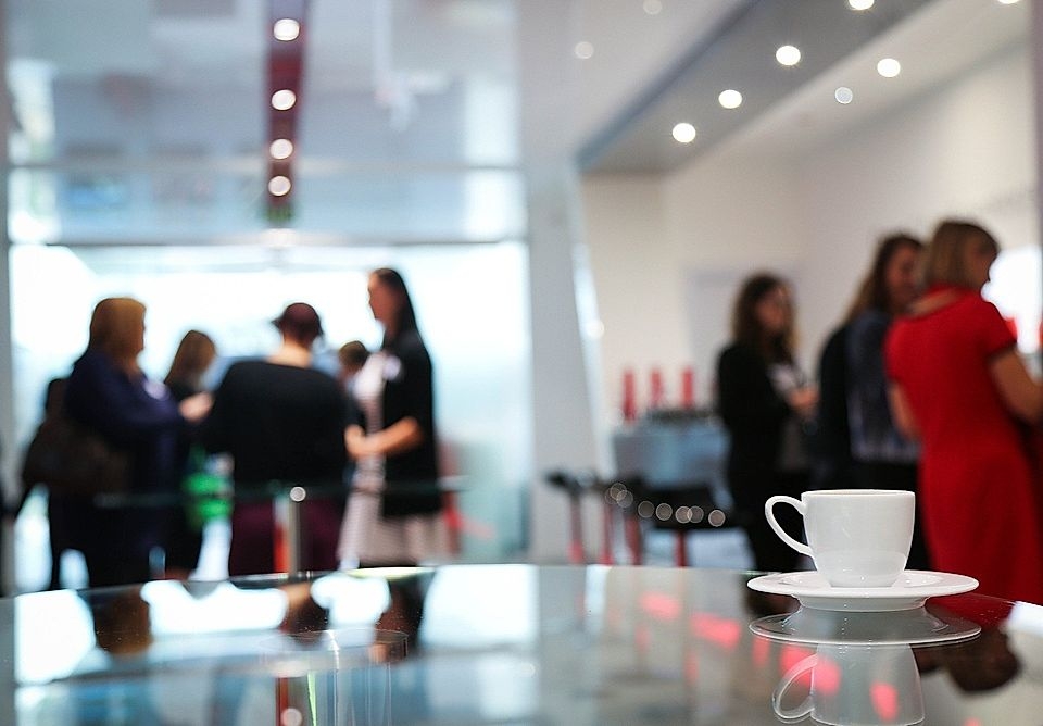 A coffee cup on a glass table in a blurred business networking event, with professionals chatting in the background.
