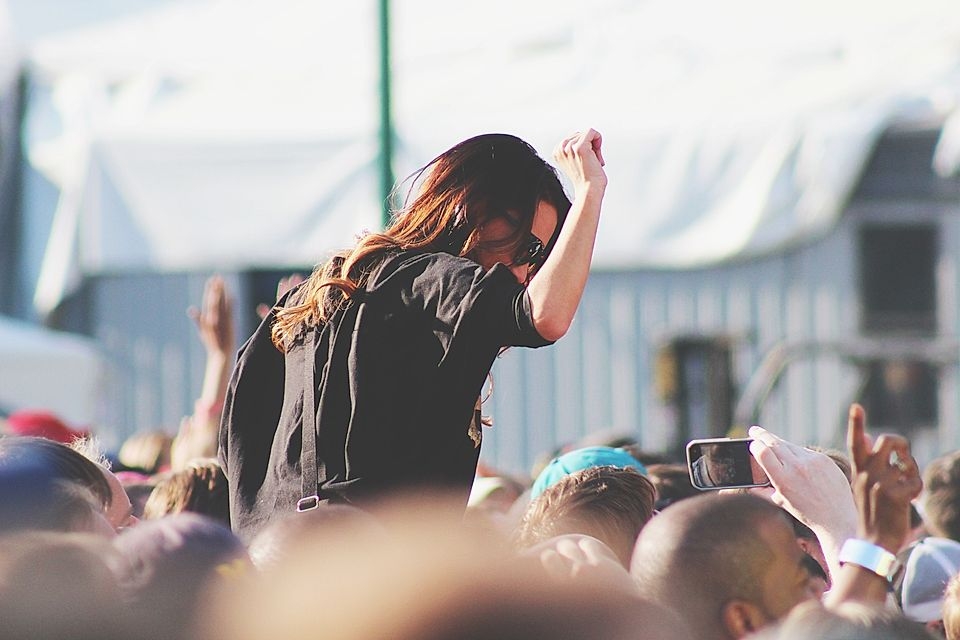 A woman with long hair dances on someone's shoulders at a crowded outdoor concert, surrounded by cheering fans.