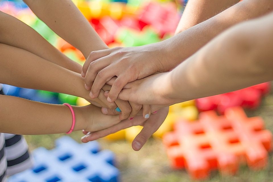 A group of hands stacked together outdoors, with colorful building blocks in the background.