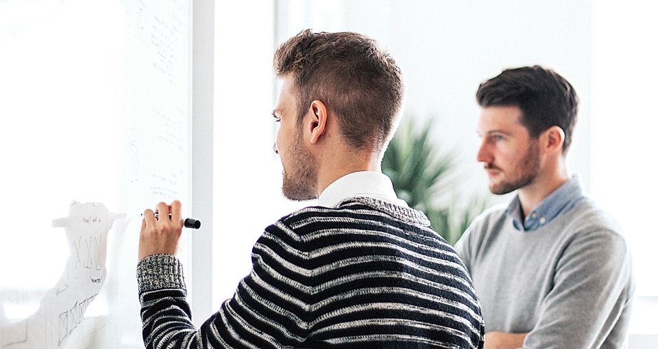 Two men collaborate in a bright office, one writing on a whiteboard while the other observes thoughtfully.