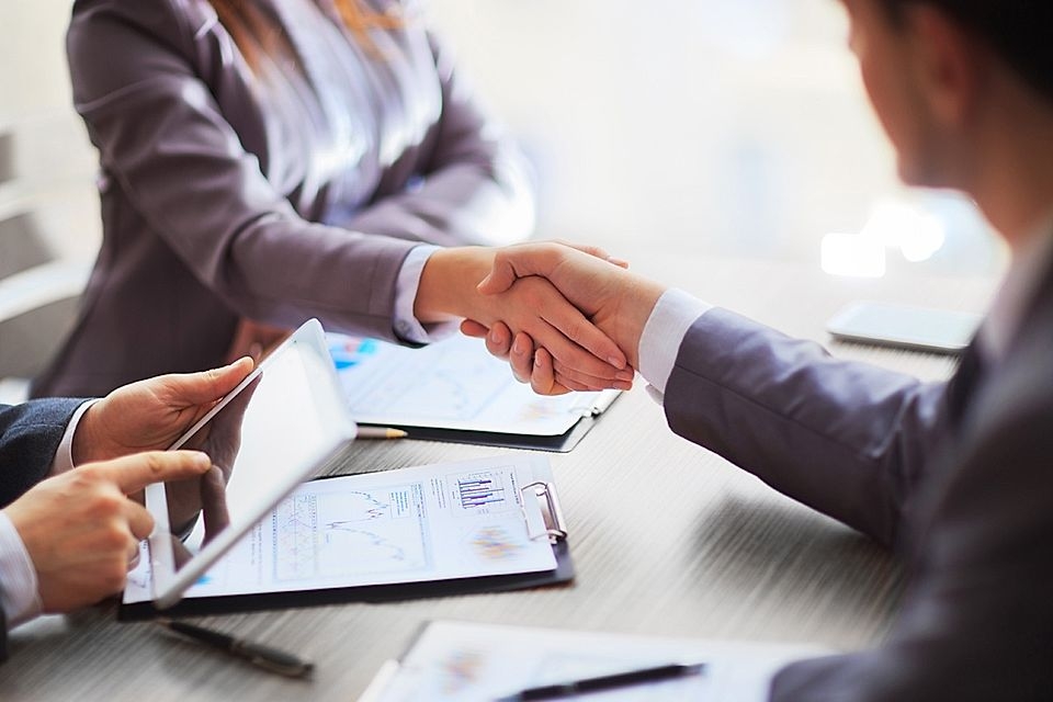Two business professionals shaking hands over a desk with documents and a tablet, symbolizing a partnership.