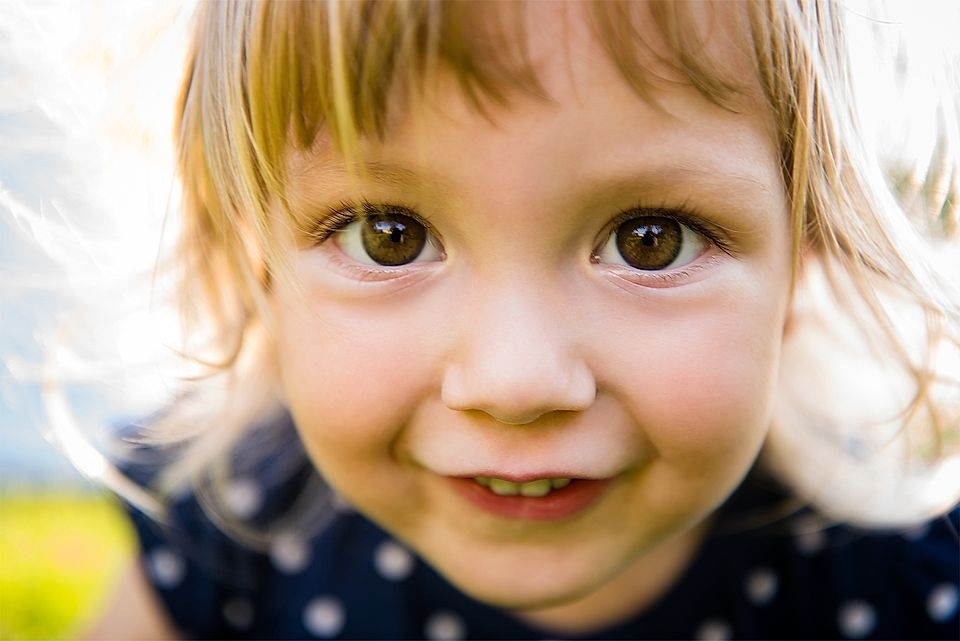 Close-up of a smiling child with brown eyes and light brown hair, wearing a polka-dot dress in a sunny outdoor setting.