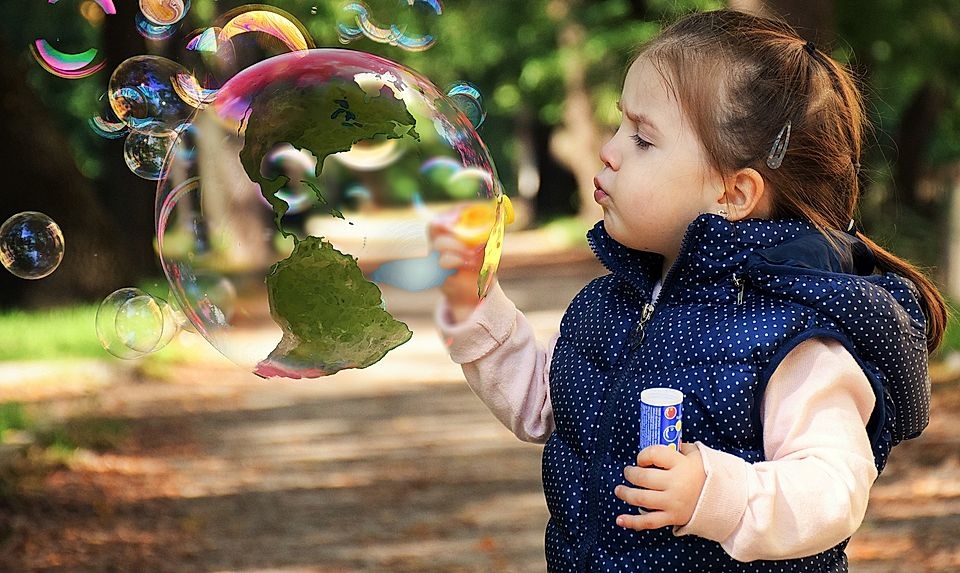 A young girl in a blue vest blows bubbles with a world map visible inside one bubble in a park setting.