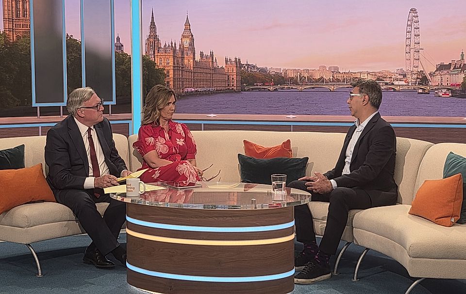 TV talk show set with three people seated on a sofa, London skyline backdrop, and glass coffee table.