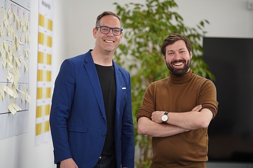 Two smiling men standing in an office beside a wall covered with sticky notes