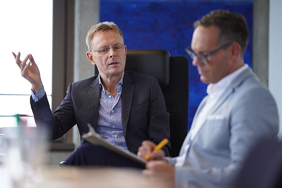 Two businessmen in a meeting, one gesturing while the other takes notes