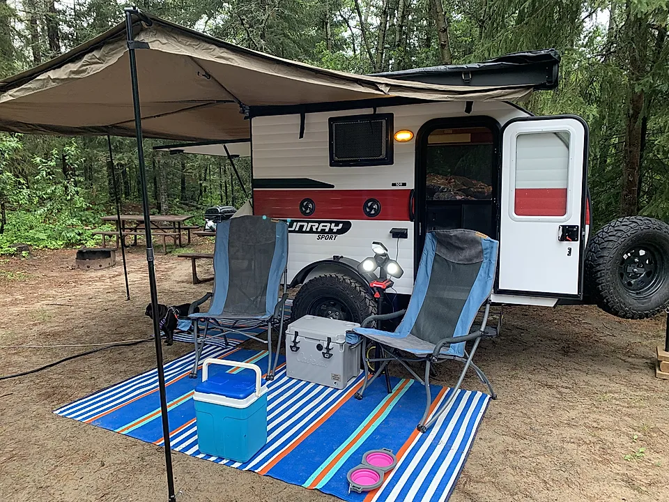 Camper trailer setup at a forest campsite with awning, chairs, cooler, and picnic table.