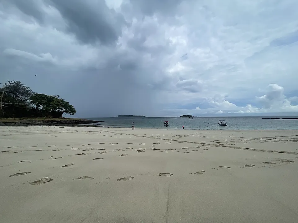 Cloudy beach scene with footprints in the sand, distant boats, and a few people near the water