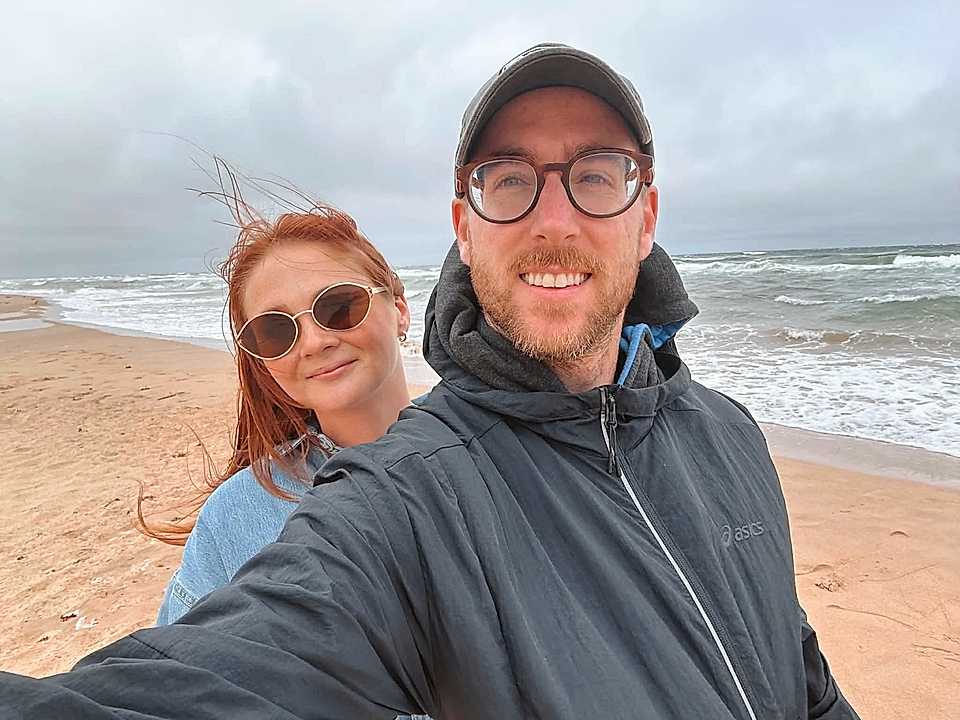 Two people taking a selfie on a windy beach with ocean waves in the background