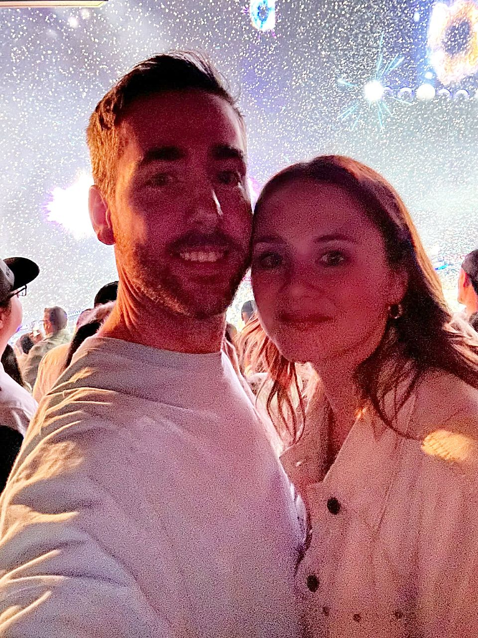 Selfie of a couple in a crowded concert or event with colorful lights overhead
