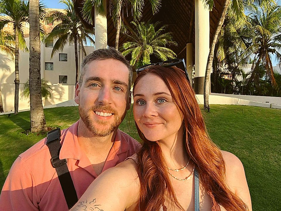 Selfie of a smiling couple outdoors with palm trees and resort buildings in the background