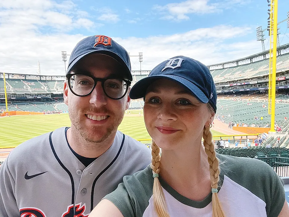 Two people taking a selfie at a baseball stadium with field and stands in the background.