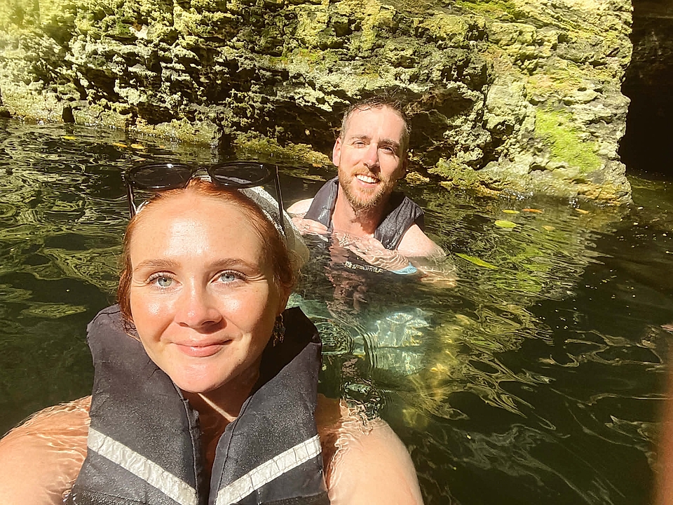 Two people in life jackets swimming in clear water near a rocky cave wall