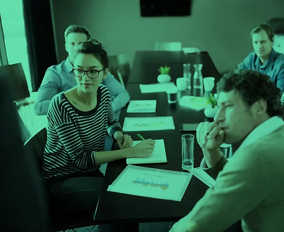 Meeting setting with five individuals seated around a conference table, engaged in discussion; documents and glasses of water visible.