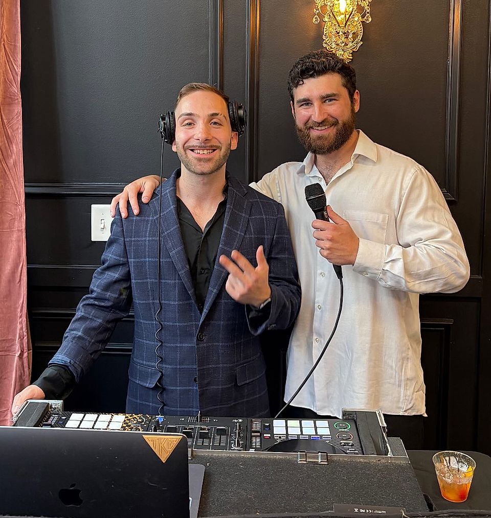 Two men at a DJ booth, one holding a microphone beside a laptop and mixer