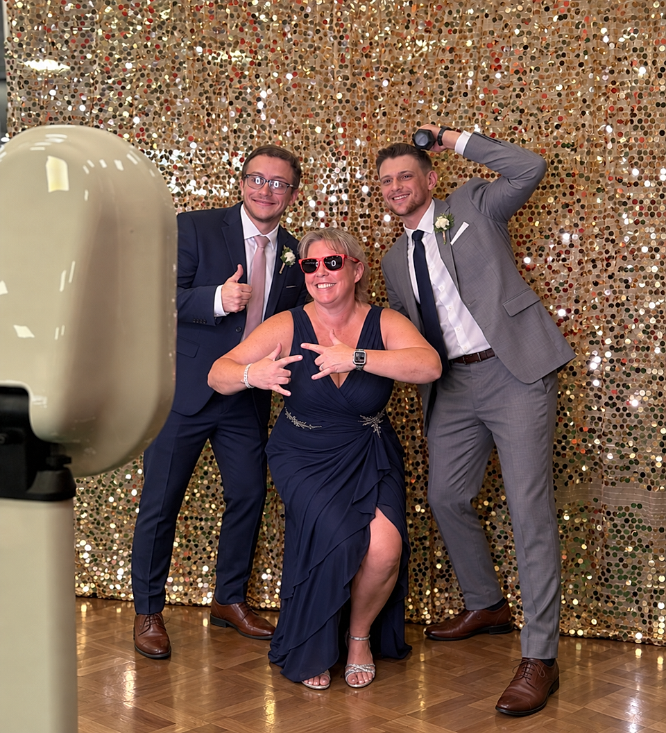 Three people posing in formal attire at a photo booth with a gold sequin backdrop.