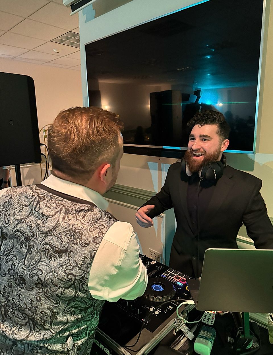 Two men talking beside a DJ setup with laptop and turntable in front of a large TV.